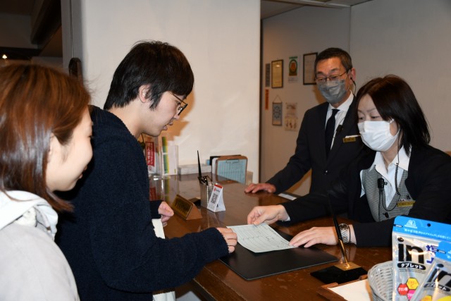 マスク姿の従業員に対し「ノーマスク」で受け付けをする宿泊客（左）＝伊東市吉田