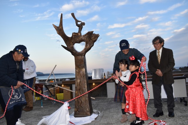 彫刻家重岡建治さん（右）とともにモニュメントを除幕し完成を祝う子どもたち＝東伊豆町の北川温泉ねこさい広場（２７日）