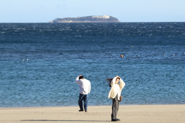 風が強い日が続き、潮でかすむ初島と風でなびく髪を押さえる人＝熱海市多賀地区の長浜海浜公園（１７日）