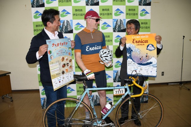 ビンテージ自転車の前でポスターを見せ合う「名誉村長」の山下市長（右）と大会関係者＝伊豆の国市役所